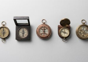 Minimalist flat lay photograph of five different antique compasses and navigation tools arranged neatly on a clean light grey textured surface, each tool slightly different in design representing different approaches to finding direction, soft natural side lighting, neutral cool color palette, no text, no people, professional editorial product photography, metaphor for choosing the right method among several valid options, 16:9 wide format